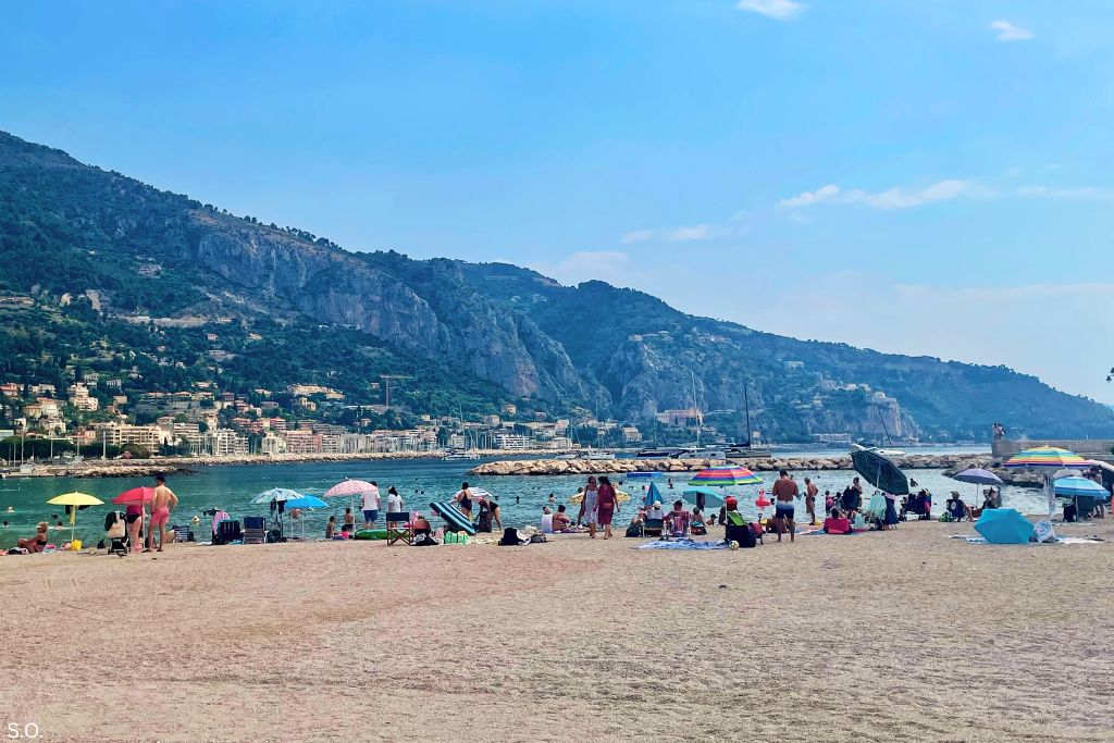 Plage des Sablettes in Menton, a crescent-shaped beach with crystal-clear waters, framed by pastel-colored buildings and the Mediterranean Sea